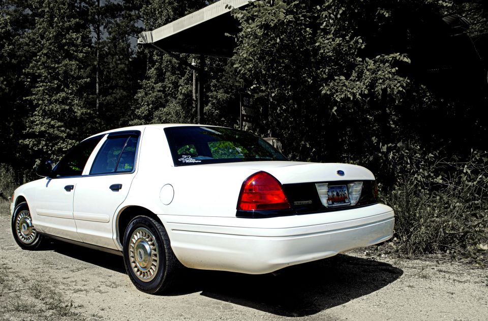 White 2002 Ford Crown Victoria parked on dirt road, trees and overhang in background
