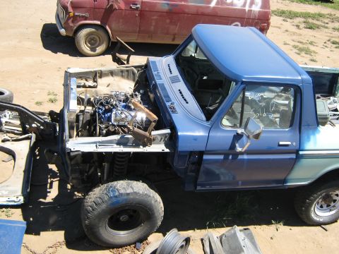 1978 Ford Bronco - A blue pickup truck with its hood open, revealing the engine, sits in a junkyard next to a red truck.