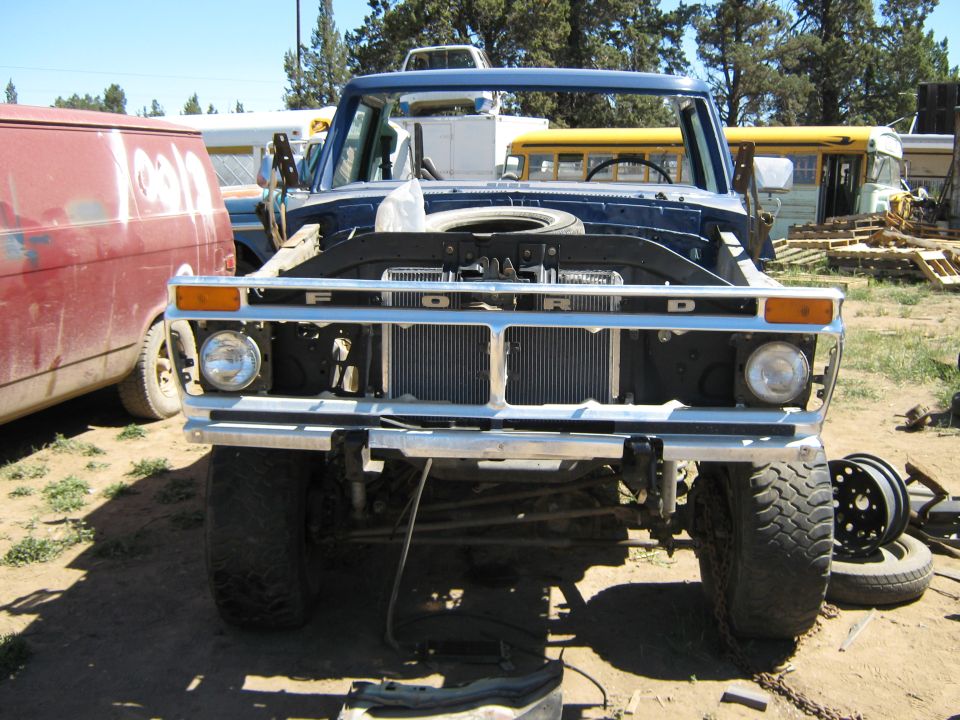 Front view of a blue Ford truck with hood removed, parked next to a red van and school buses
