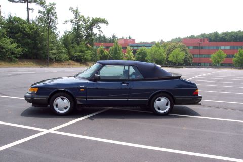 2001 Ford Crown Victoria - A dark blue Saab 900 convertible is parked in a large, empty parking lot with a red brick building in the background.