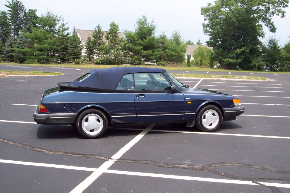Blue convertible parked in an empty lot with trees and houses in the background