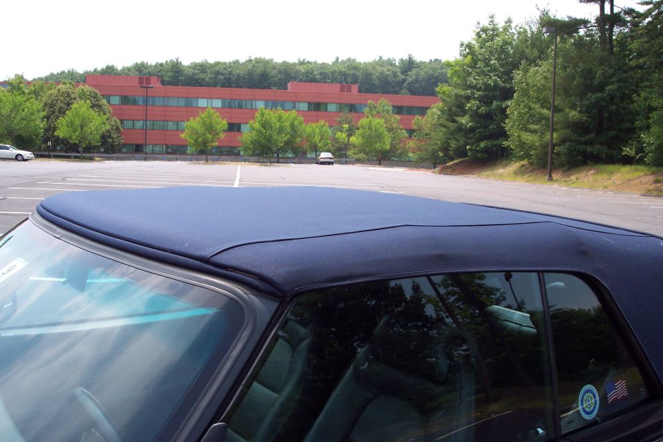 Car's blue convertible top and interior, with a red brick building and trees in the background. Another car is parked in the distance