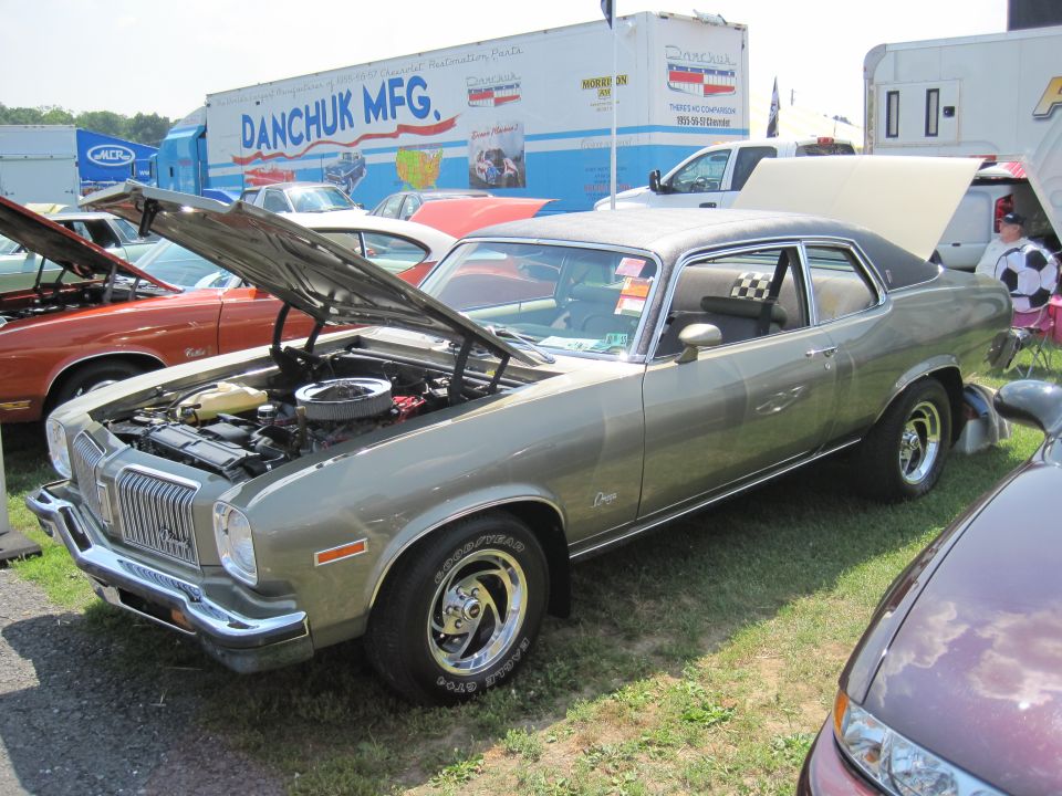 1975 Oldsmobile Omega coupe with hood open, parked on grass at an outdoor event. Another car is visible in the foreground