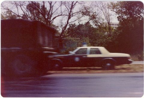 2001 Ford Crown Victoria - A police car is seen in motion next to a large truck, with trees and a building in the background.
