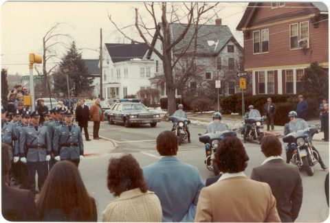 2001 Ford Crown Victoria - A police escort, motorcycles and a sedan, moves through a residential street lined with onlookers and houses.