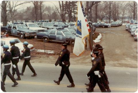 2001 Ford Crown Victoria - A procession of police officers marches past numerous police vehicles parked in a lot.
