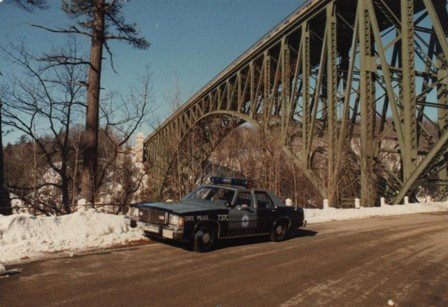 2001 Ford Crown Victoria - A police car is parked on the side of a road with snow on the ground, beneath a large metal bridge.