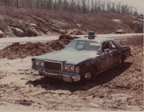 2001 Ford Crown Victoria - A dark police car, marked "STATE POLICE 796", is parked on a dirt shoulder next to a road.