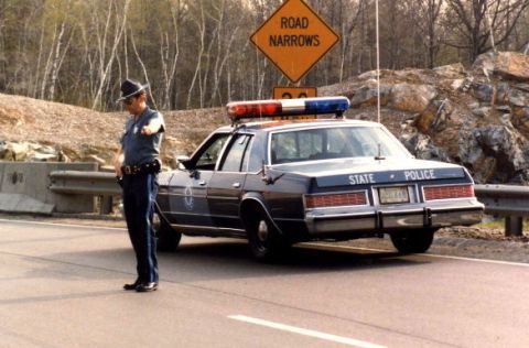 2001 Ford Crown Victoria - A state trooper directs traffic next to a marked police cruiser, with a 'Road Narrows' sign in the background.