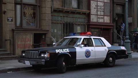 2001 Ford Crown Victoria - A police car with a light bar is parked on a city street in front of buildings with various signs.