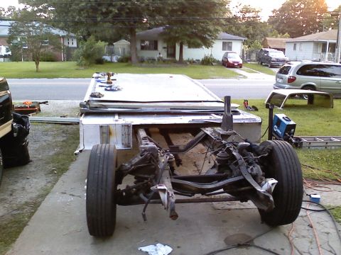 1990 Ford Bronco - Front view of a stripped truck frame on a driveway with the body and drivetrain removed, showing the bare frame rails, coil-spring front su…