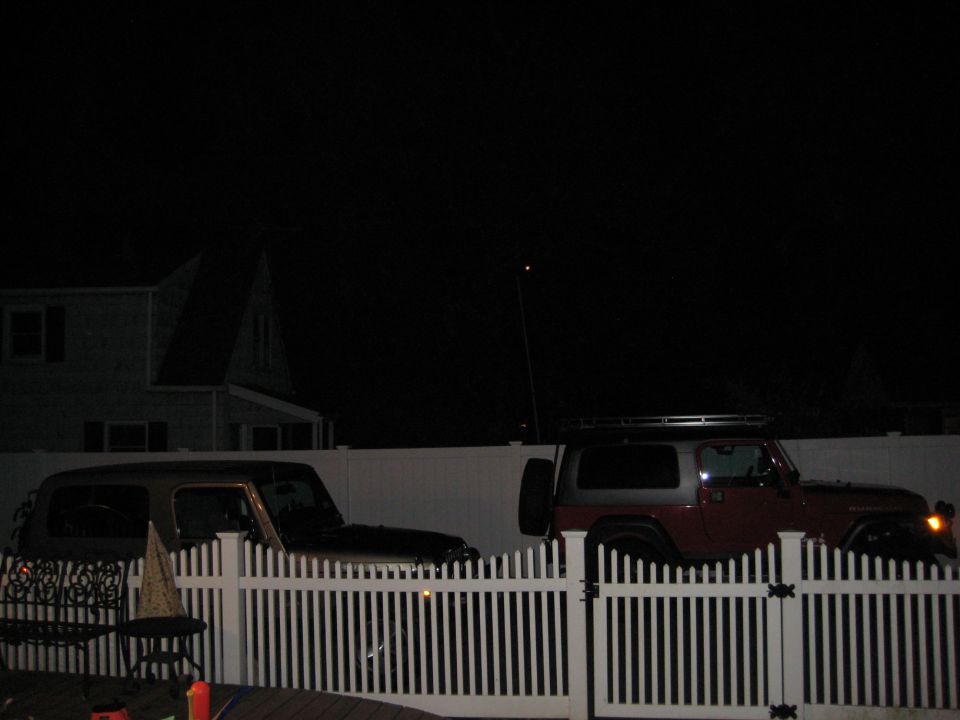Two Jeep Wranglers parked behind a white picket fence at night. One is dark colored, the other is red and silver with a roof rack