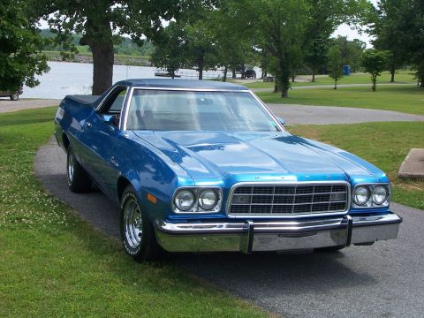 1973 Ford Ranchero - A blue 1973 Ford Ranchero is seen from a three-quarter front view, parked on a paved path next to a grassy area and a lake.