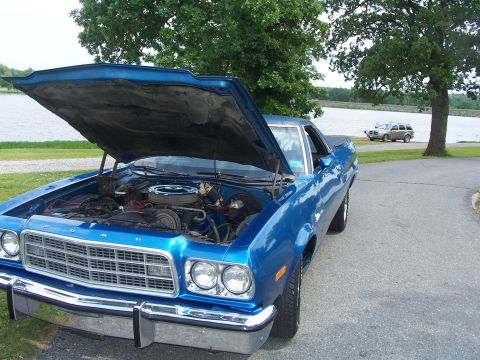 1973 Ford Ranchero - A blue 1973 Ford Ranchero is shown with its hood open, parked near a body of water with another vehicle in the background.
