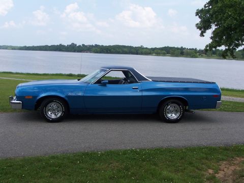 1973 Ford Ranchero - A blue 1973 Ford Ranchero is parked on a road next to a large body of water under a cloudy sky.