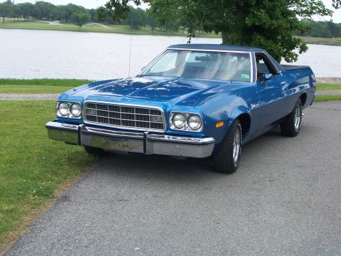 1973 Ford Ranchero - This blue pickup truck is parked on a paved surface next to a grassy area and a lake.