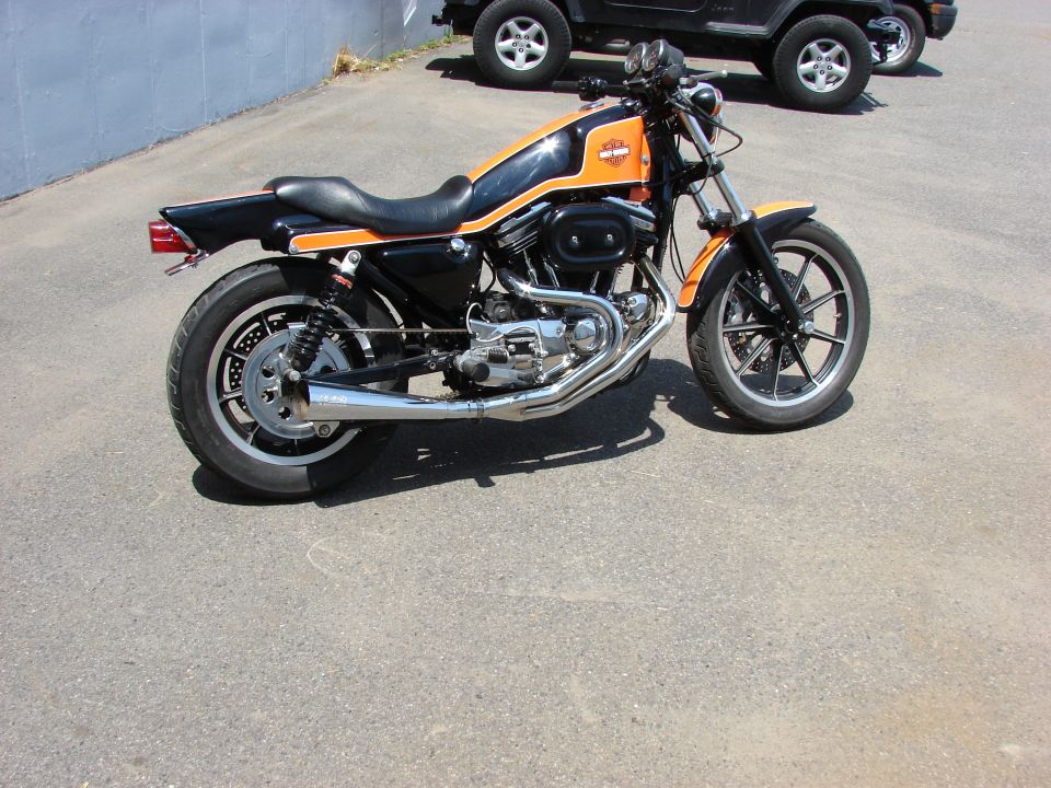 Harley-Davidson Sportster motorcycle with orange and black paint, parked near two SUVs