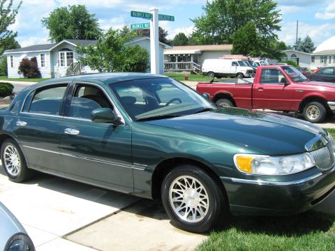 1988 Mercury Grand Marquis - A green sedan is parked in front of houses, with a red pickup truck and white van visible behind it.
