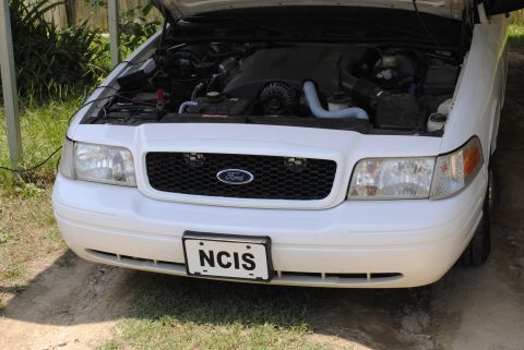 2002 Ford Crown Victoria - Front view of a white Ford Crown Victoria with the hood up, revealing the engine compartment and a custom NCIS license plate.