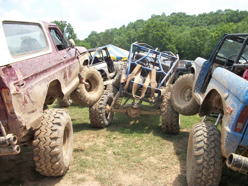 Mud-covered lifted off-road trucks and a tube buggy parked on grass at an outdoor event