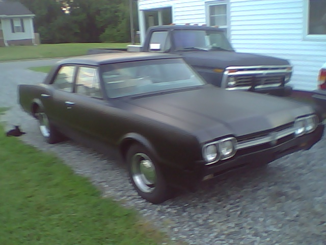 Flat black 1960s two-door coupe with quad headlights on a gravel driveway
