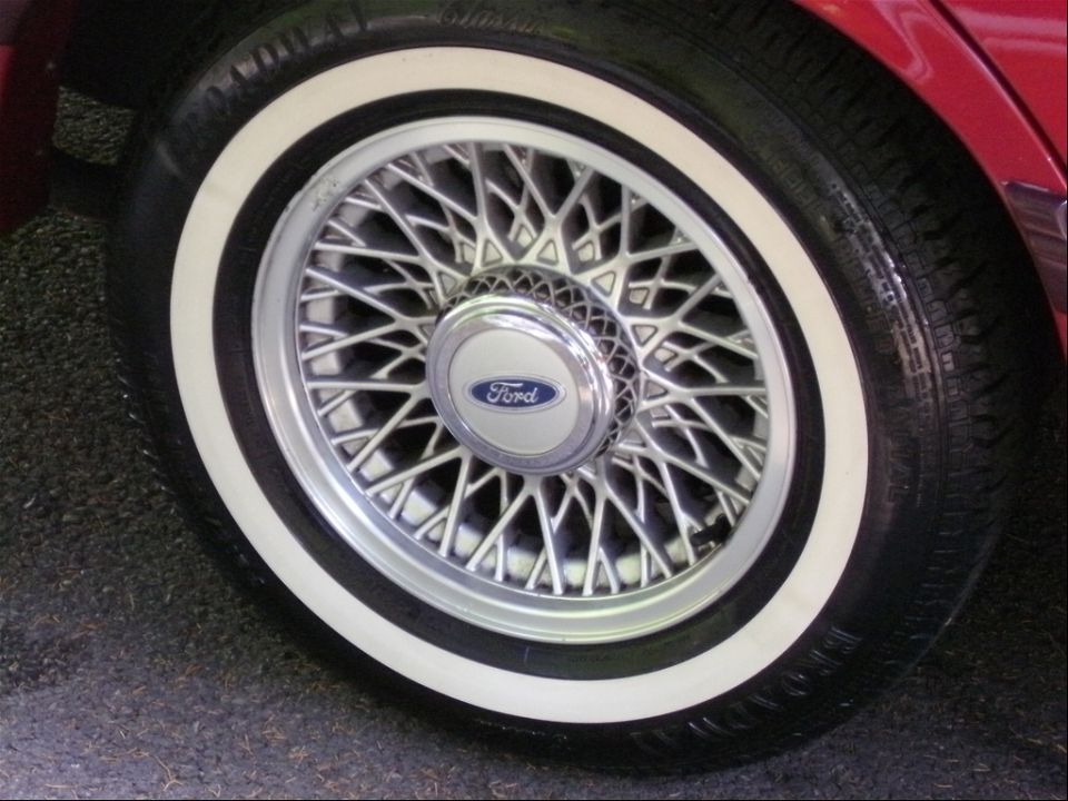 Silver lace-spoke alloy wheel with Ford center cap and whitewall Broadway Classic tire on a red car