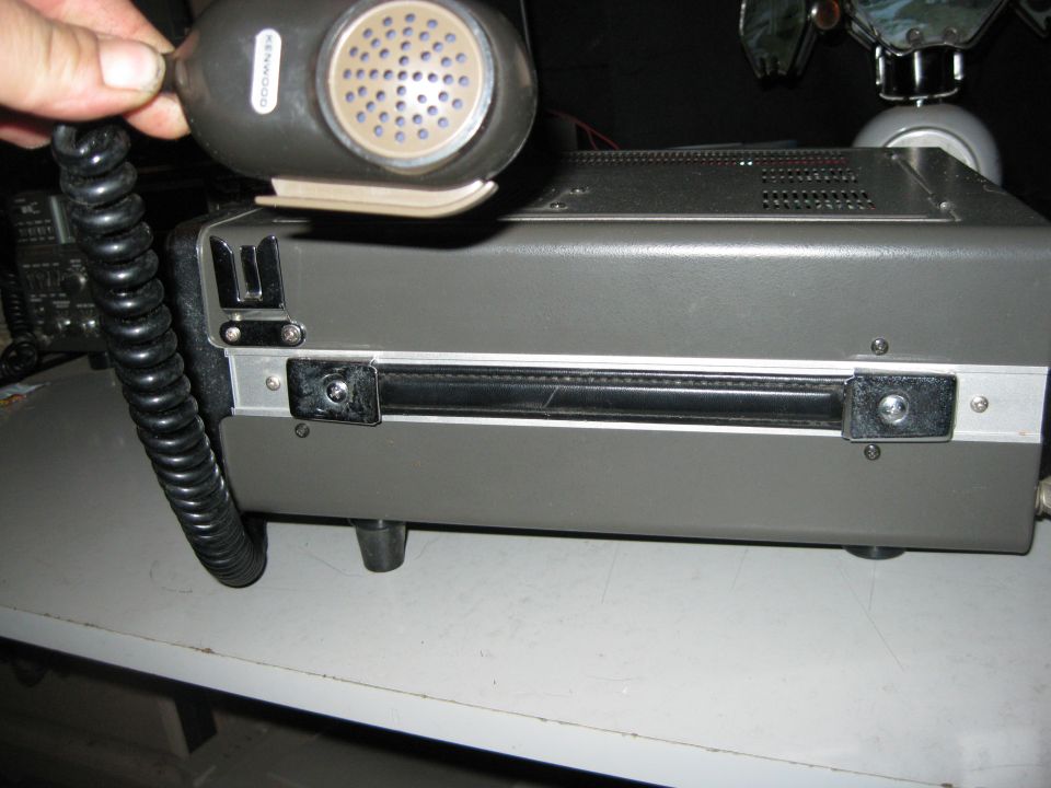 Gray radio transceiver with black coiled cord microphone held by a hand. The radio has a silver trim and a black handle