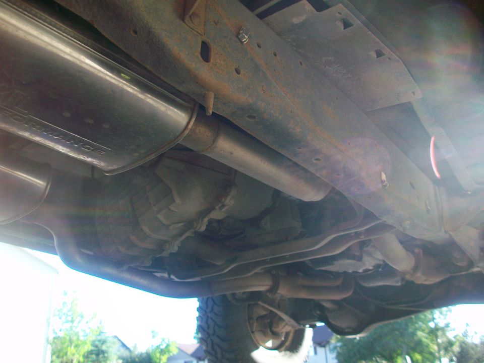 Underside view of vehicle frame, exhaust system, and transmission. Some rust visible on the frame