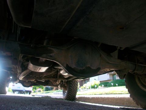 1986 Ford Bronco - A low-angle view of a vehicle's undercarriage, revealing mechanical components and a glimpse of the surrounding environment.