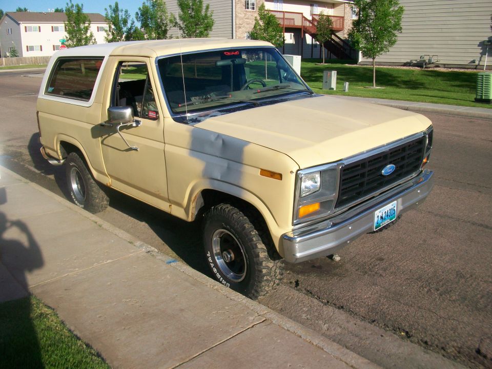 Tan Ford Bronco parked on a residential street, showing front and side views. The vehicle has a white removable top and a dent on the hood