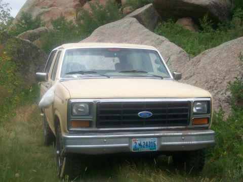 1986 Ford Bronco - A tan 1986 Ford Bronco is seen from the front, parked amidst large rocks and grassy vegetation.
