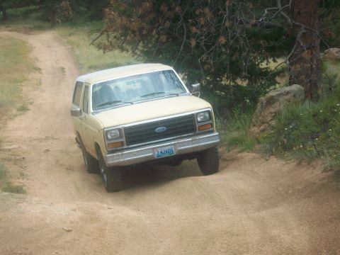 1986 Ford Bronco - This tan Ford Bronco navigates a dusty, off-road path with trees and rocks in the background.