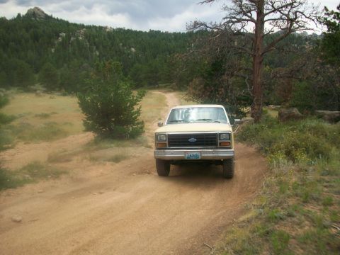 1986 Ford Bronco - A tan 1986 Ford Bronco navigates a dirt road through a wooded, hilly landscape.