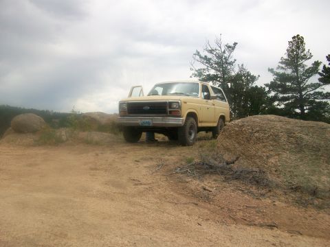 1986 Ford Bronco - A yellow Ford Bronco is parked on a dirt path with large rocks and trees in the background under a cloudy sky.
