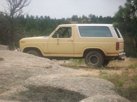 1986 Ford Bronco - A yellow Ford Bronco is seen from the side, parked on a rocky trail with trees in the background.