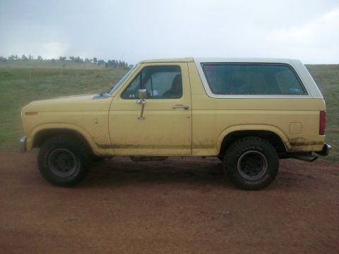 1986 Ford Bronco - A yellow 1986 Ford Bronco with a white hardtop is parked on a dirt road.
