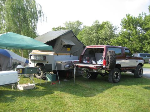 1996 Ford Bronco - A red and silver SUV tows a pop-up camper trailer, set up for camping with tents and a grill visible.