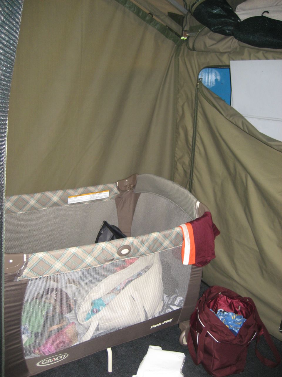 Inside a tent, a playpen is filled with baby items. A maroon backpack sits nearby