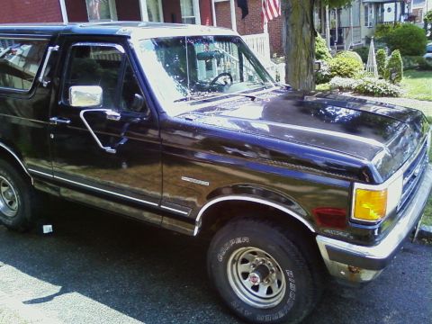 1990 Ford Bronco - Sunlit 3/4 front view of a dark blue two-door full-size Ford Bronco parked at a curb in front of brick row houses, showing chrome window tr…