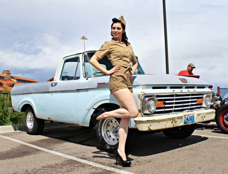 Woman in a vintage military-style outfit poses by a light blue Ford pickup truck with a chrome grille
