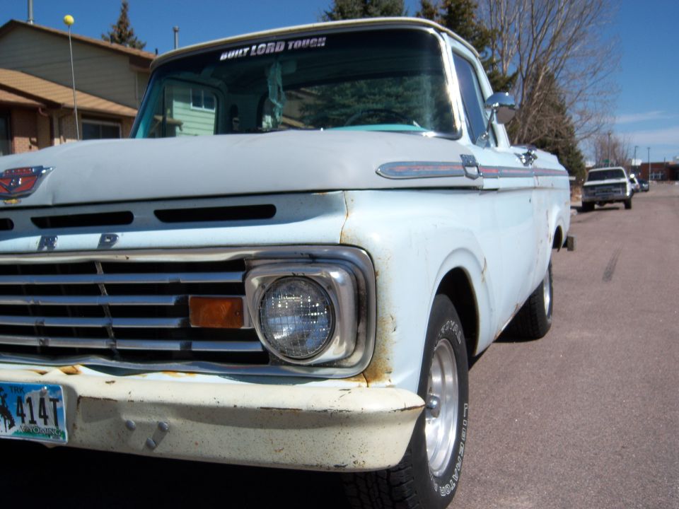 1963 Ford F-100 pickup truck front quarter view, light blue paint, chrome grille, and bumper. Another white truck is visible in the background