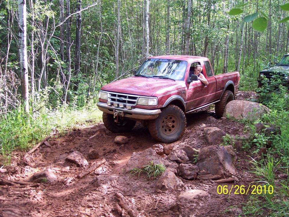 Red pickup truck driving on a muddy, rocky trail in the woods. Another vehicle is visible in the background