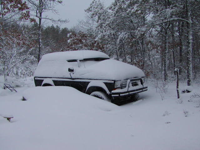 Dark SUV buried in deep snow among snowy trees, front three-quarter view