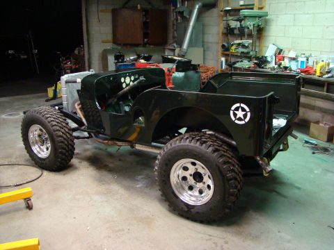 1948 Jeep Wrangler - A dark green Jeep-style vehicle build in mid-restoration, shown from a 3/4 front-right angle in a workshop, with a white military-style sta…