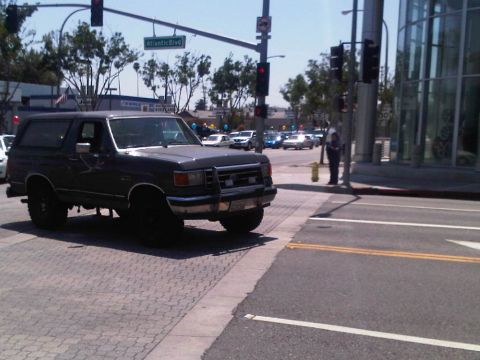 1986 Ford Bronco - A dark SUV drives through an intersection on a sunny day, with other vehicles visible in the background.