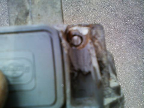 1990 Ford Bronco - Blurry macro shot of a cast-metal module's mounting ear held over a concrete driveway, showing a corroded bolt hole and heavy white powdery…