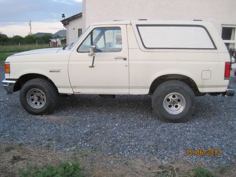1989 Ford Bronco - A white Ford Bronco is parked on a gravel surface next to a building. The vehicle is viewed from the side.