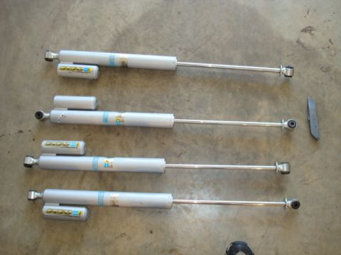 1995 Ford Bronco - Four Bilstein shock absorbers are displayed on a concrete surface, alongside a detached metal bracket.