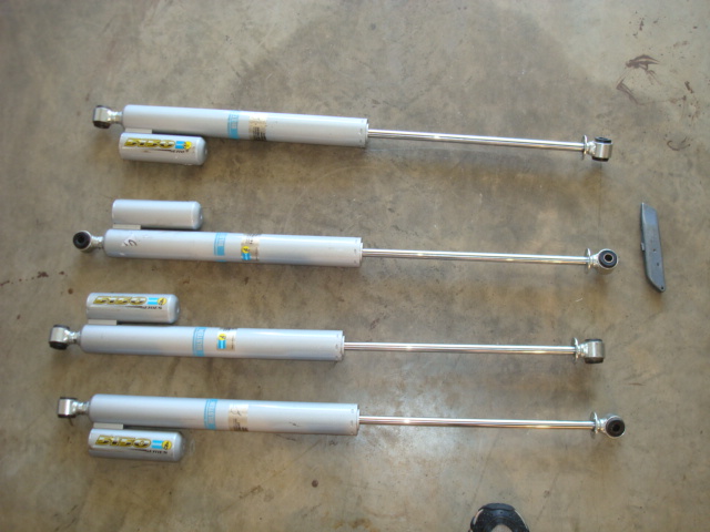 Four silver shock absorbers with yellow and blue labels, laid out on a concrete floor next to a gray bracket