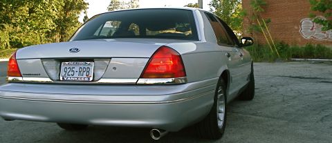 2000 Ford Crown Victoria - The rear of a silver sedan is shown parked outdoors with trees and a brick wall in the background.
